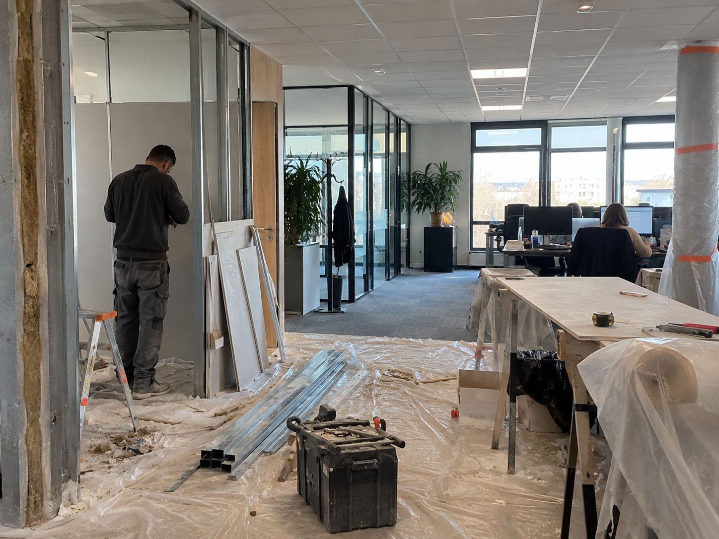Travaux de cloisonnement dans des bureaux en activité, zone protégée et postes de travail occupés en arrière-plan.